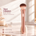Glimmer rose-gold brush vibrator with two-button controls, shown on an ivory vanity beside pearls and a candle.